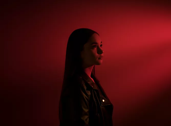 A profile portrait of a woman with long, dark hair, wearing a dark leather jacket. The image is dominated by a deep, monochromatic red light that illuminates the subject's face and creates a distinct spotlight effect against the red background, leaving the rest of her in silhouette.