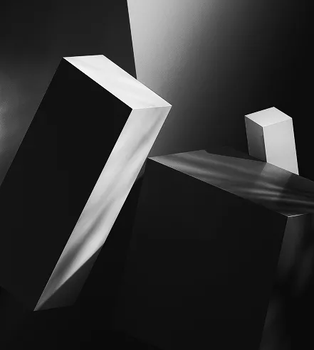 Abstract black-and-white composition featuring tall angular geometric blocks lit by dramatic light, creating sharp contrasts and deep shadows.