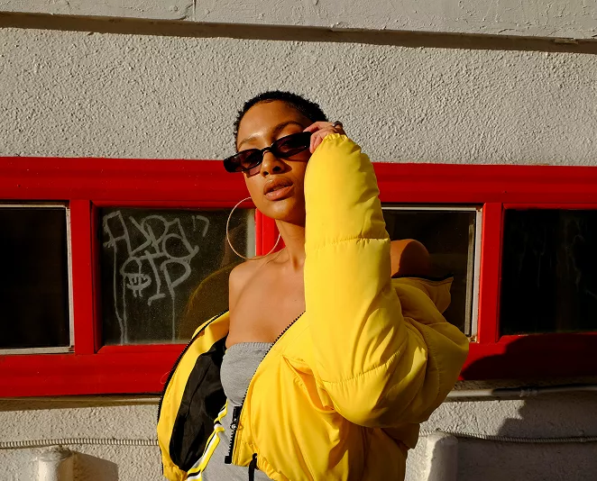 Person in yellow puffer jacket adjusting small sunglasses while standing outdoors against red-trimmed windows with graffiti in sunlight.