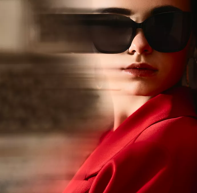 A stylish woman wearing large dark sunglasses and a red outfit stands in soft light as motion blur sweeps across the frame, creating a dramatic modern look.