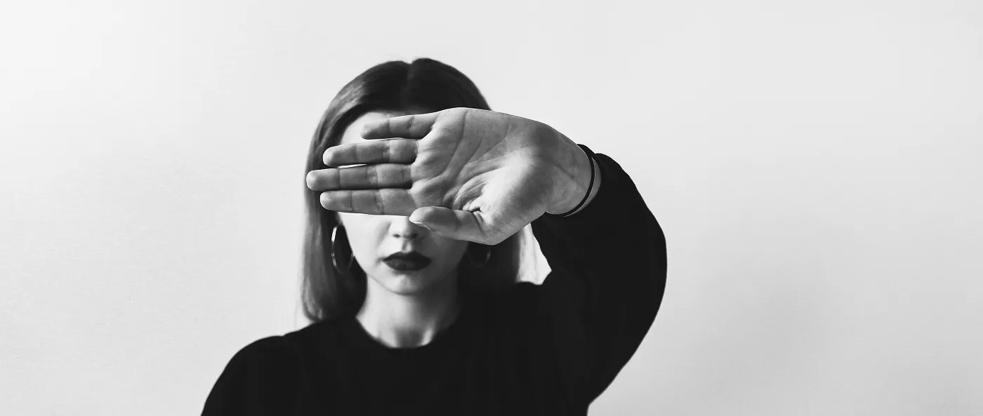 A black and white, close-up shot of a woman covering her face with one hand, palm forward, in a gesture that suggests a refusal to be seen or to face the viewer. She is wearing hoop earrings and dark lipstick.