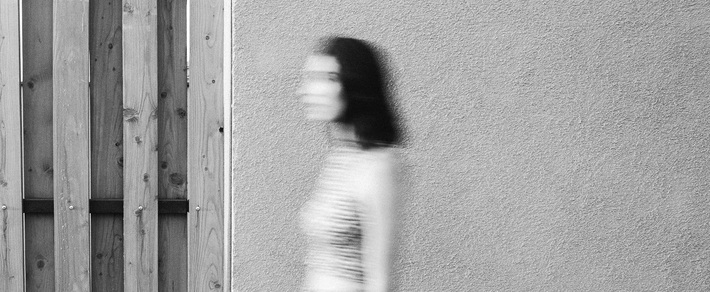A blurred woman in a striped top walks past a textured wall beside wooden panels, creating a motion-blur effect in an otherwise still, minimal scene.