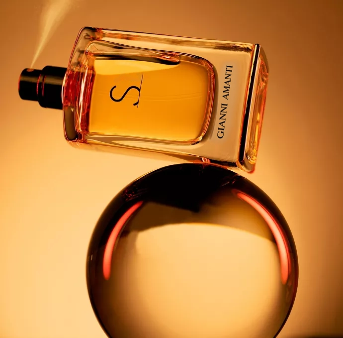 The bottle is positioned diagonally and appears to be balanced on a large, reflective, circular object . The scene is illuminated by warm, orange/amber light, and a fine mist of spray is visible near the top of the bottle.